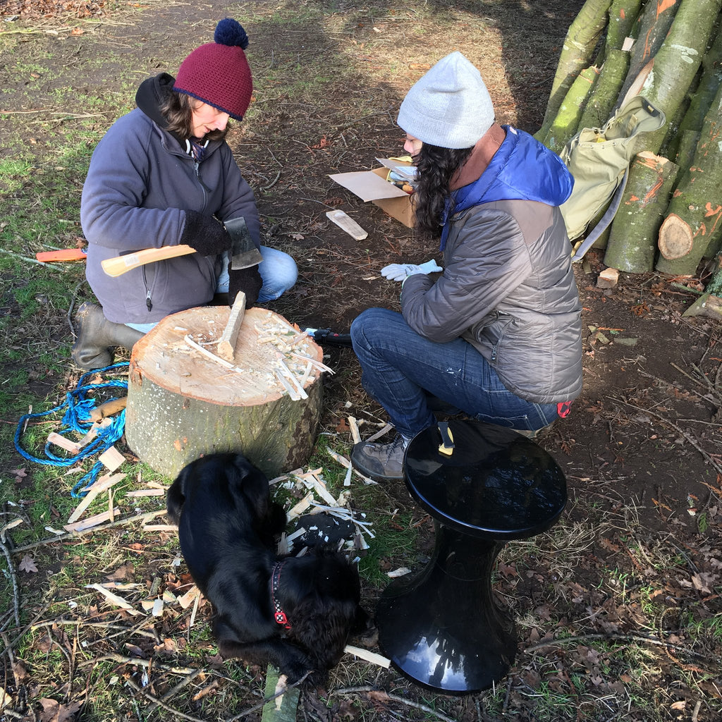 Photo of two people sitting outside, one (left) chipping at a piece of wood, the other (right) supervising.