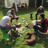 Photo of four people sitting outside on tree stumps, carving spoons.
