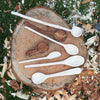 Photo of seven freshly carved spoons on top of a tree stump surrounded by wood chips.